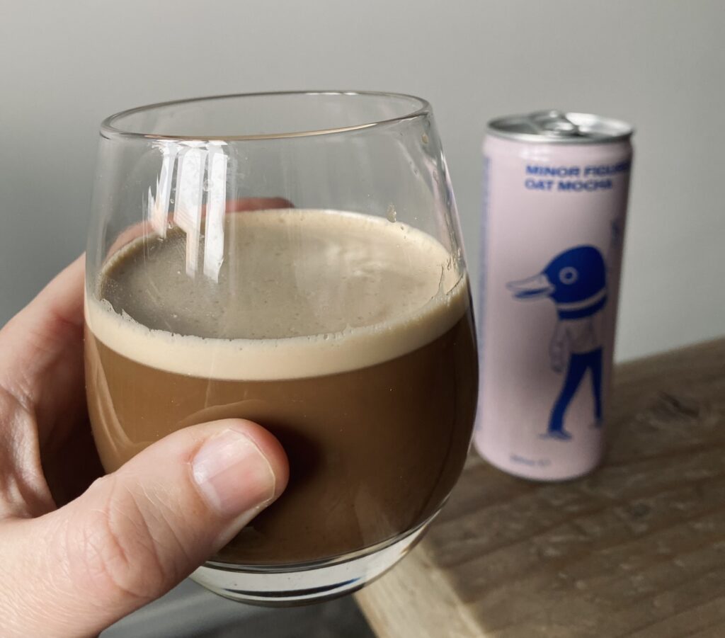 A glass of Minor Figures cold brew coffee held up in front of the camera with the can in the background