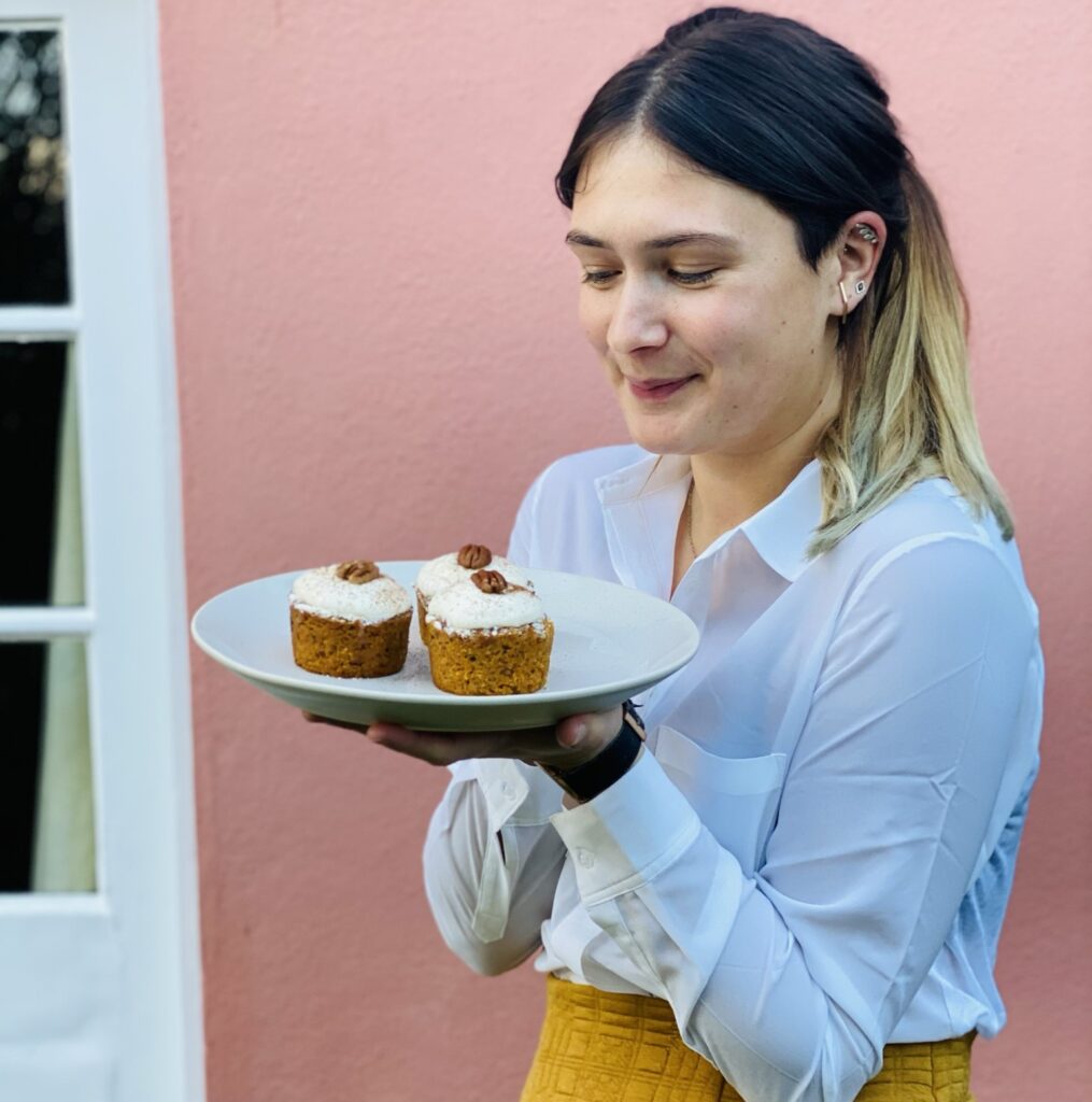 Lucy holding a plate of vegan carrot cake muffins outside in front of a pink wall