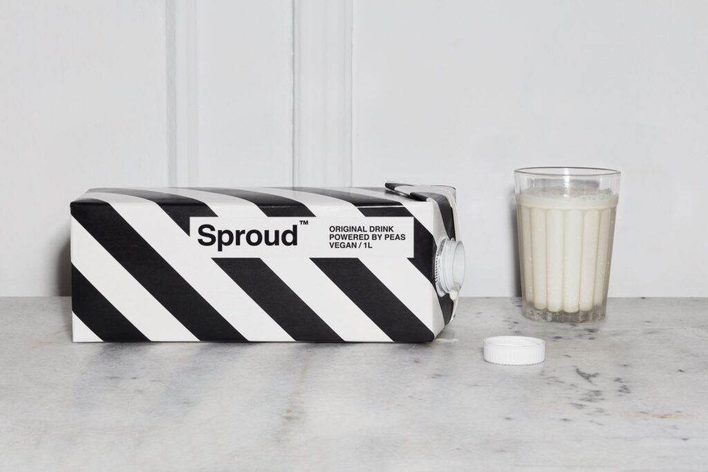 A carton of Sproud plant-based milk open and on its side pouring out pea milk next to a glass of vegan milk