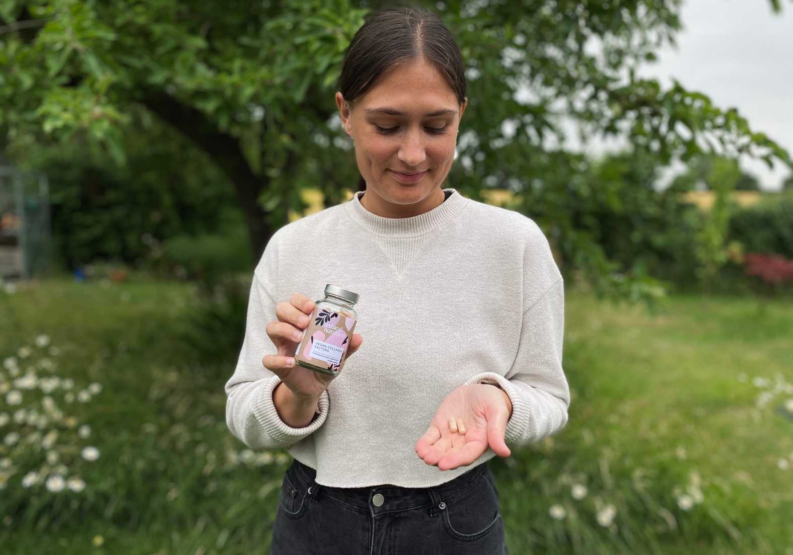 Lucy holding a jar of Ethical Nutrition vegan collagen supplements with two capsules in her other hand which she is looking down at whilst stood in the garden