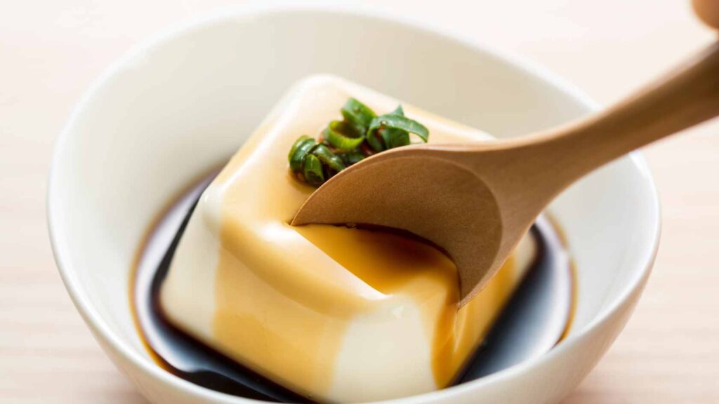 A block of silken tofu sat in a bowl of soy sauce being tucked into with a wooden spoon
