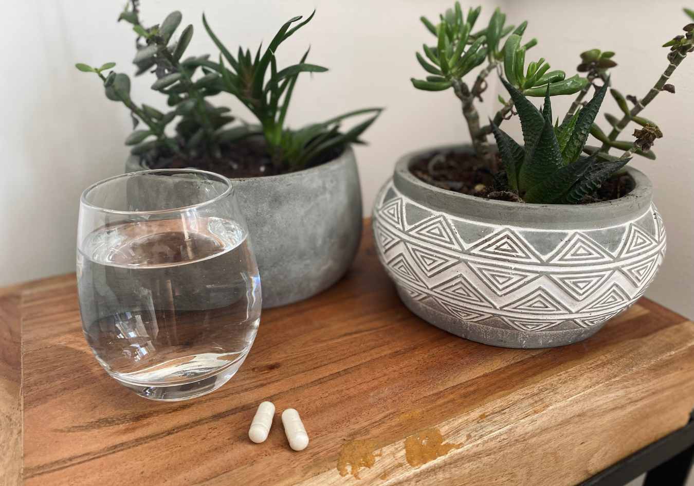 A glass of water next to two vegan vitamin B12 supplements in front of two plants on a wooden surface