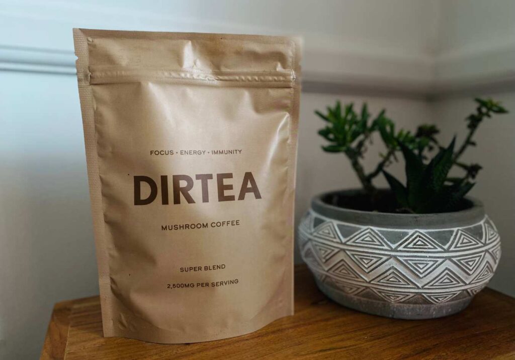 A bag of DIRTEA mushroom supplement coffee powder on a wooden surface with a plant in the background