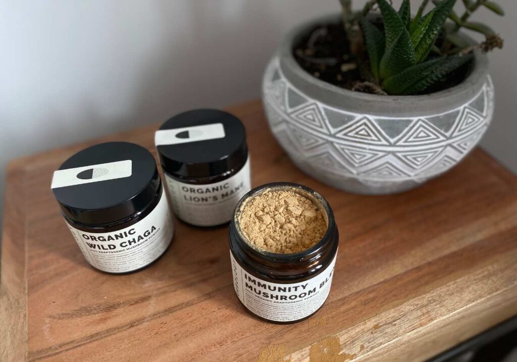 An aerial view of three mushroom powders by Erbology with one jar open showing the powder on top of a wooden surface with a plant